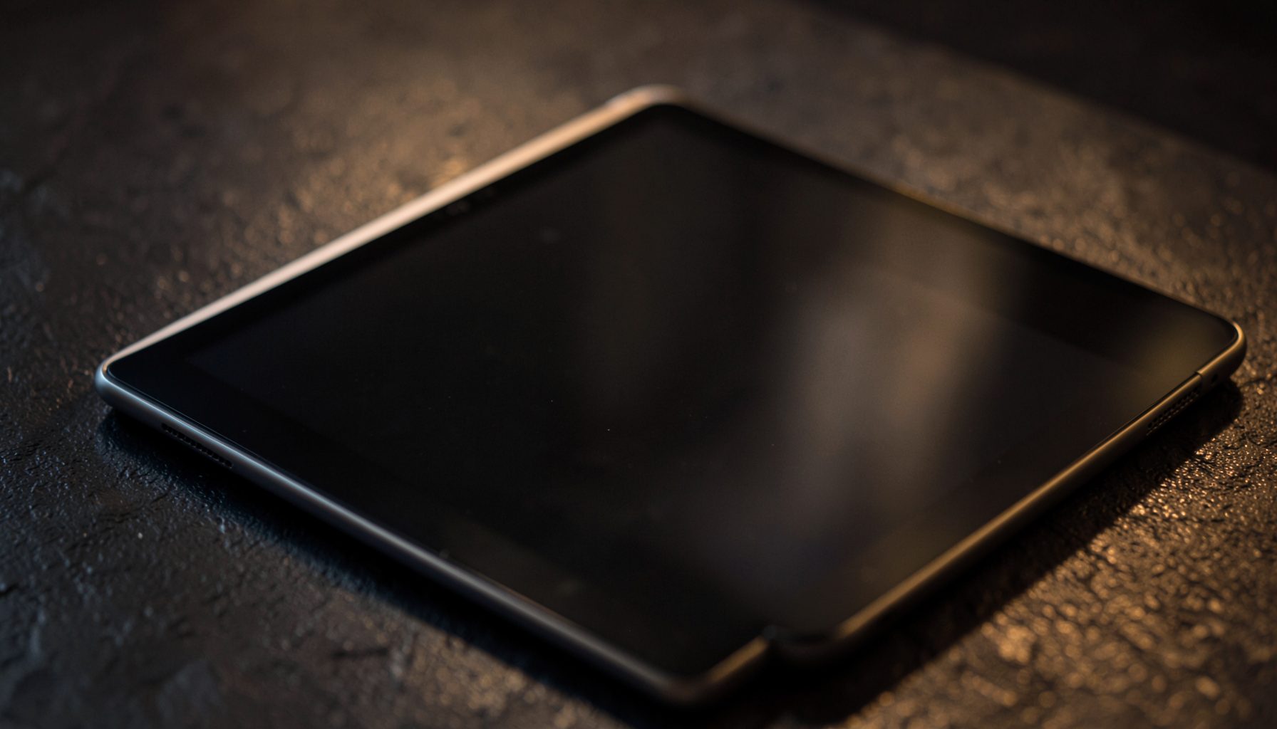 A moody close-up of a tablet and e-reader resting on a textured dark surface, with soft warm light casting gentle shadows across the scene