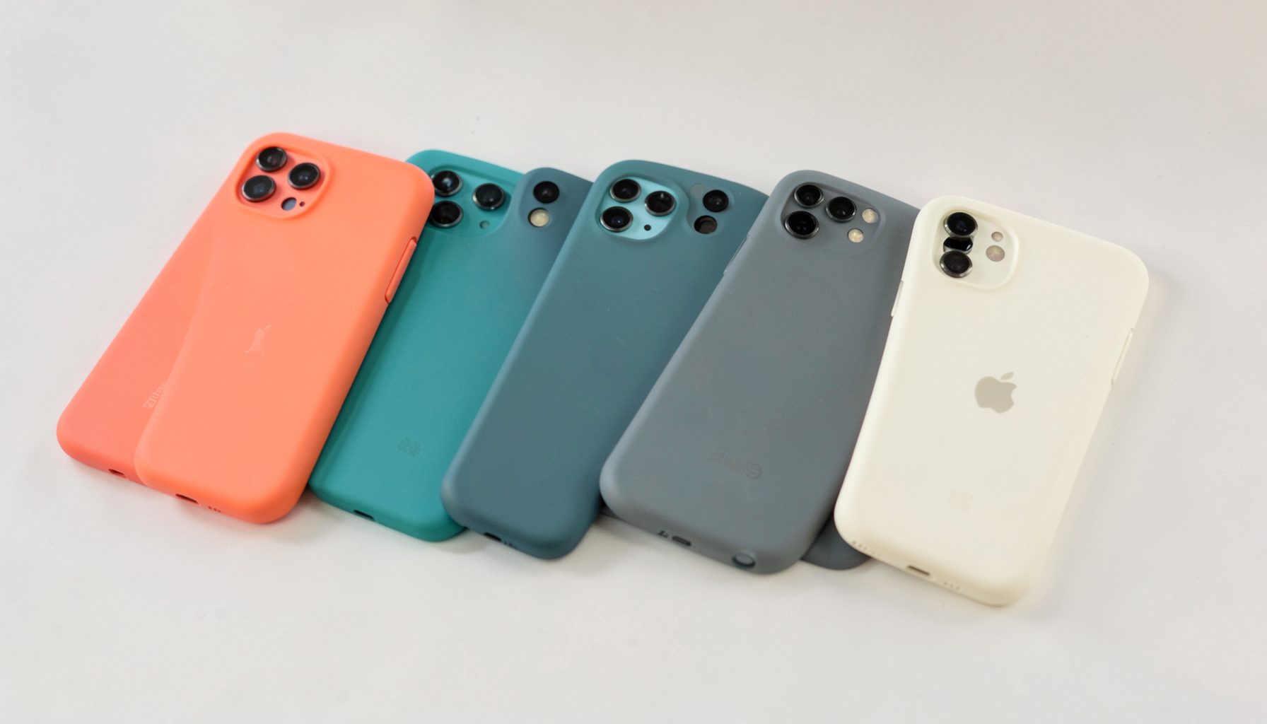 A row of colorful smartphone backs in matte finishes — coral, teal, slate, and cream — arranged diagonally on a pale neutral surface, soft daylight casting gentle shadows.