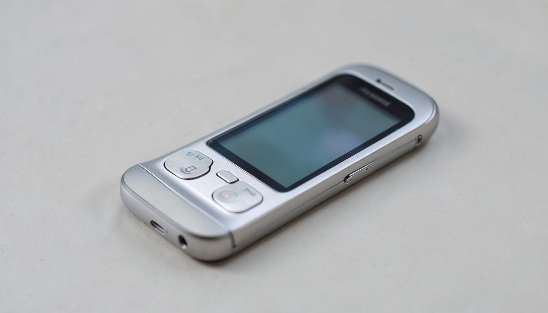 A slim silver feature phone with rounded edges and a small color display, resting on a neutral surface.